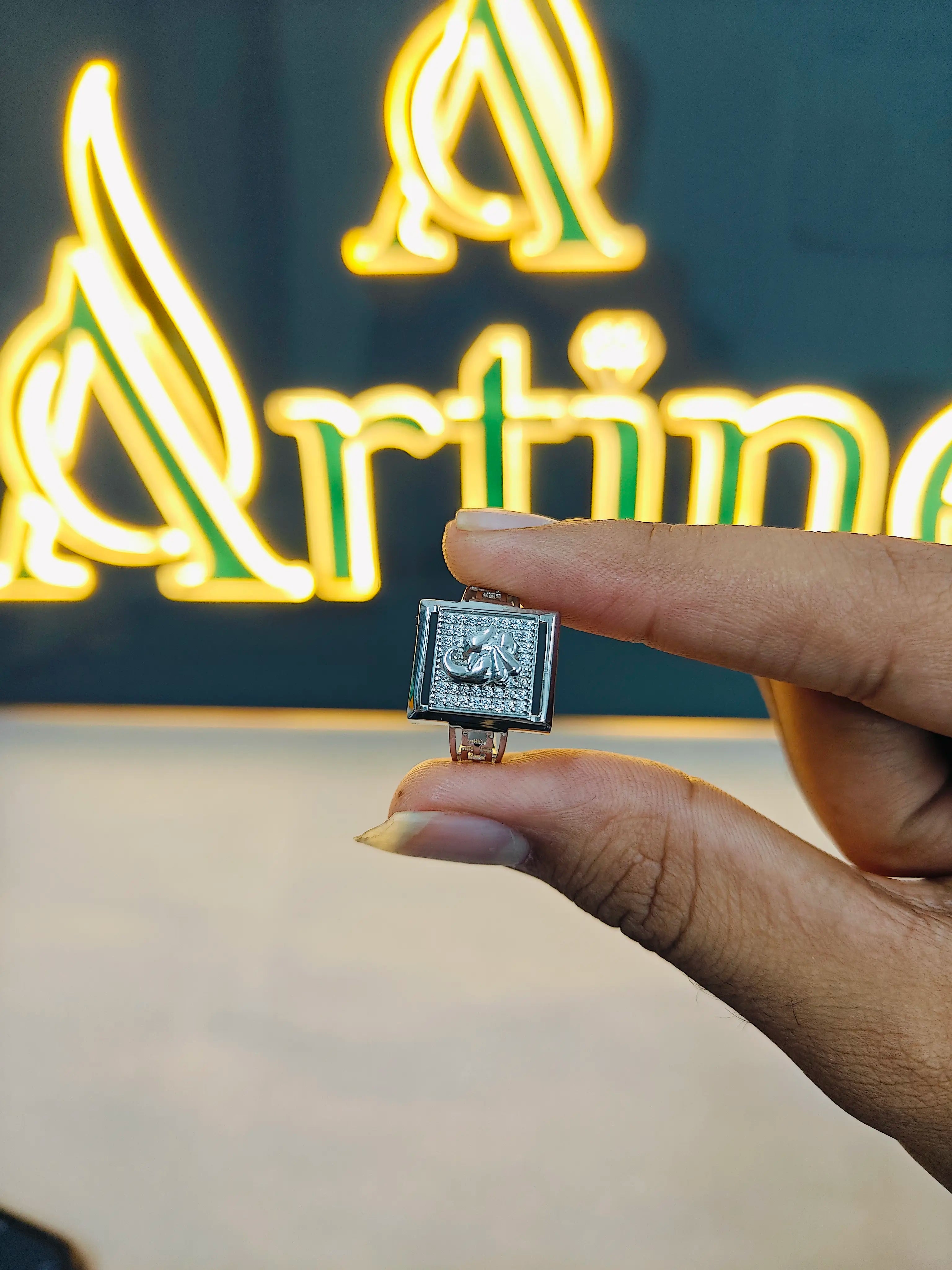 Hand holding a small silver pendant in front of a neon sign with 'Artime' branding.